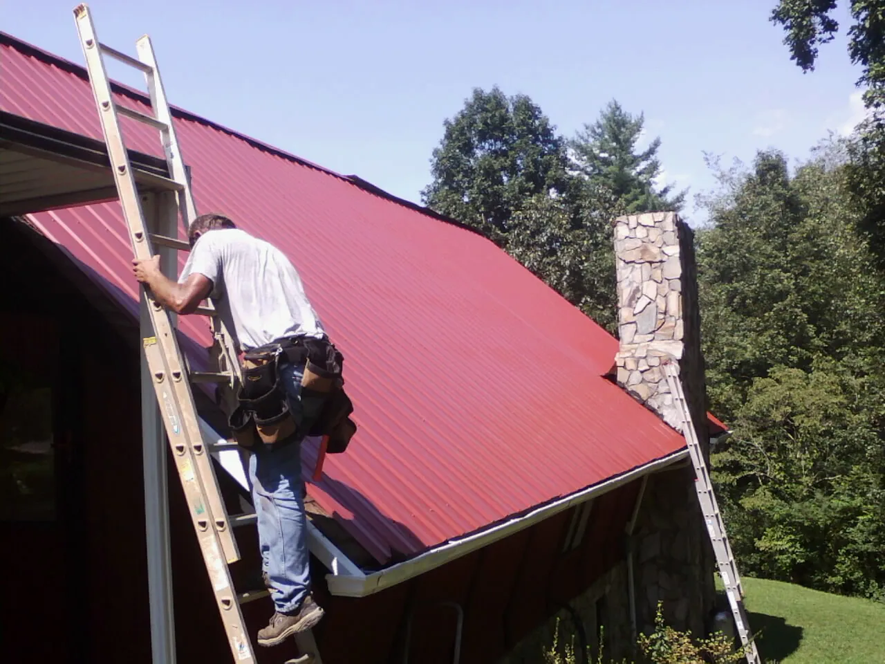 Roofer on a ladder beside a red metal roof and stone chimney — Soffit & Fascia Repair by Roofing Brothers in Searcy